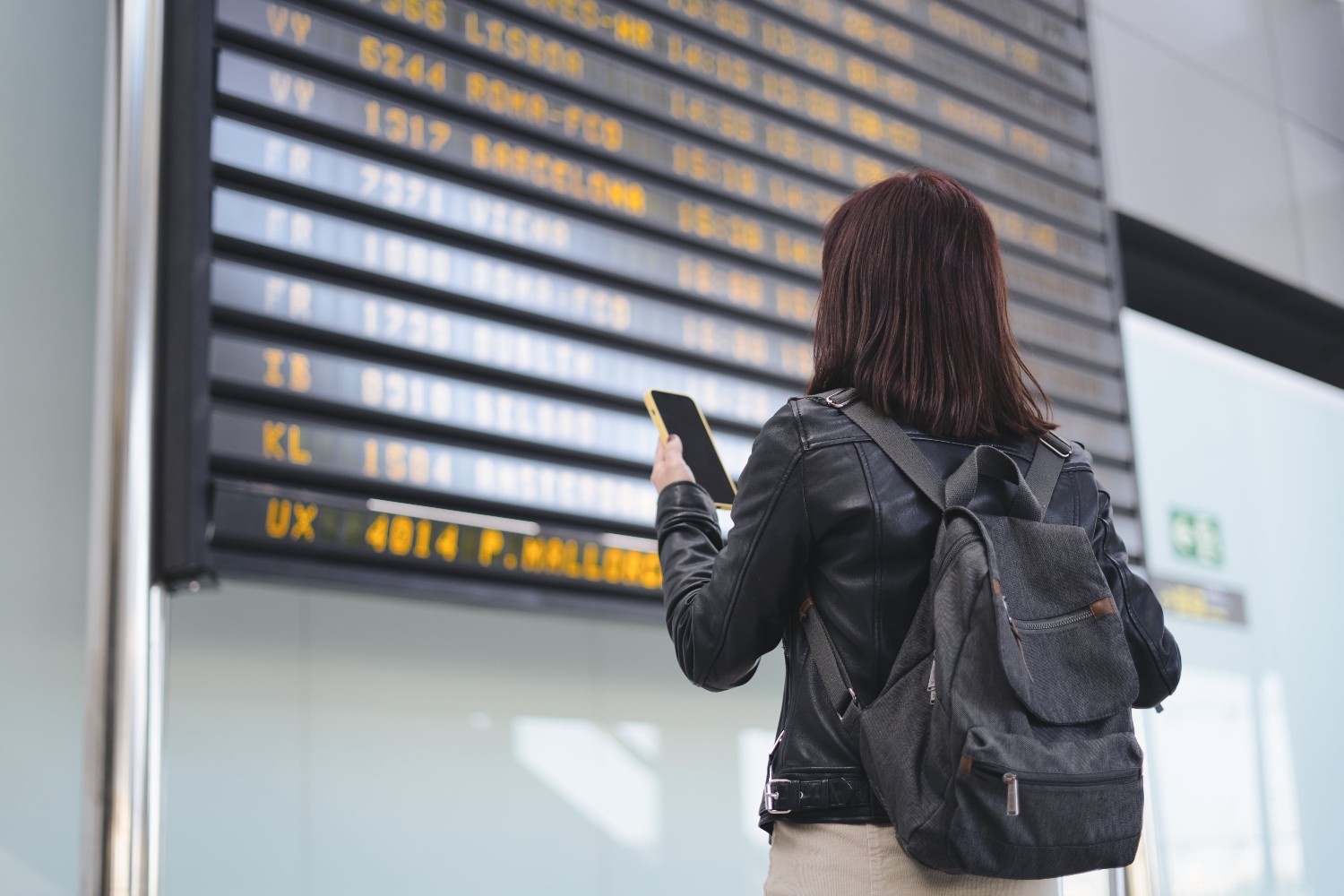 Dekle z kratkimi rjavimi lasmi s telefonom v roki gleda razpored letov na letališču